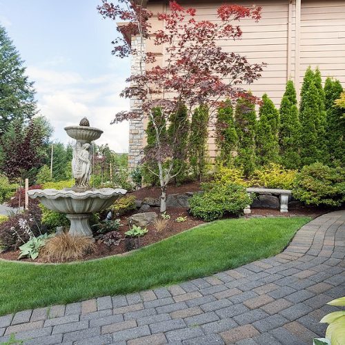 House manicured frontyard garden with water fountain stone bench green lawn plants trees shrubs and brick paver walkway path